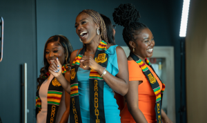 Four smiling students wearing dresses and Kente stoles stand back to back at the Black Excellence Graduation celebration