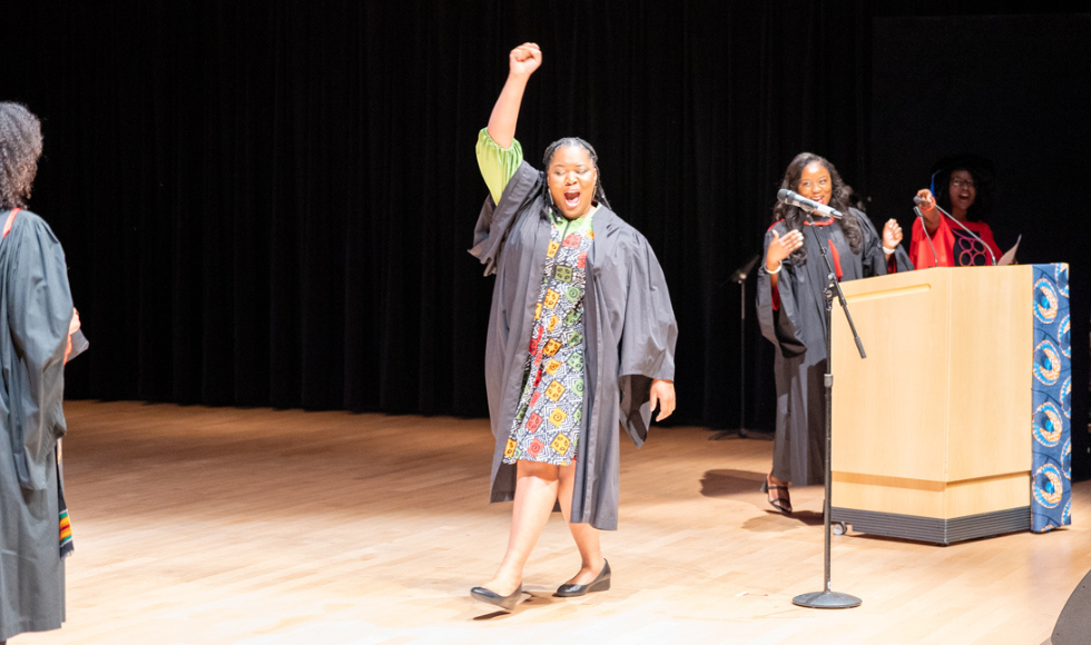 Graduating MD Kika Otiono raises her hand and whoops as she crosses the stage to receive her Kente stole at the Black Excellence Graduation celebration.
