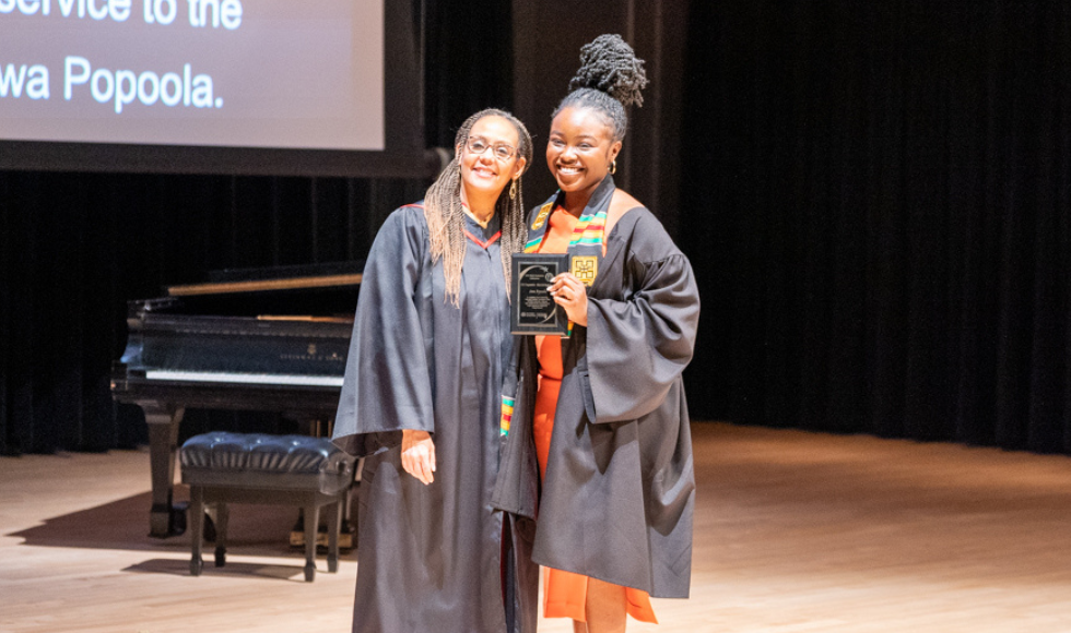 Clare Warner and Anu Popoola smile on stage while Anu holds her Black Girl Magic award at the Black Excellence Graduation celebration.