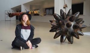 Meghan Nemeth sitting cross-legged on the floor of an art exhibition, beside an enormous spiky spherical object — a piece of art.