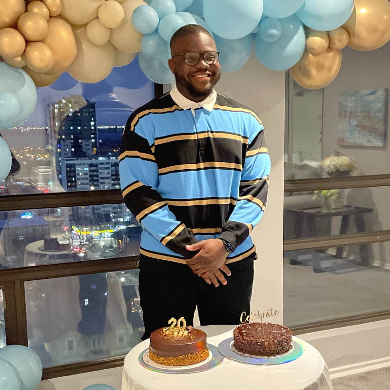 Toluwalase Dayo-Olaide standing in front of a table that has two birthday cakes on it. There are blue and yellow balloons behind him. 