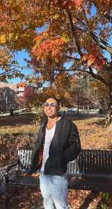 Ian Carlo wearing sunglasses on a sunny afternoon standing on campus against fall foliage