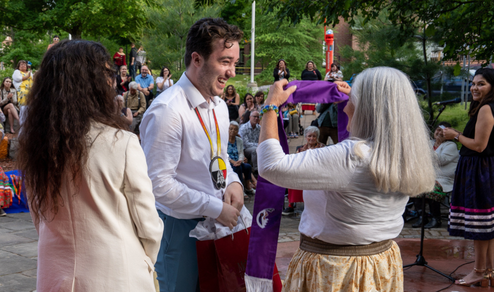 Jordan Derochie smiles at Kathie Knott [seen from behind] as she prepares to put a purple graduation stole around his neck at the outdoor classroom. The chancellor is standing nearby.