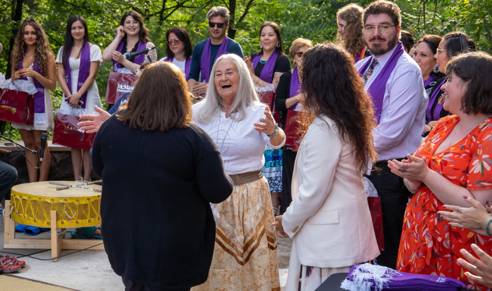 Elder Kathy Knott smiles widely and opens her arms for a hug as a graduating student (seen from behind) approaches her at the outdoor classroom to receive a graduation stole. The Chancellor is standing beside them. 