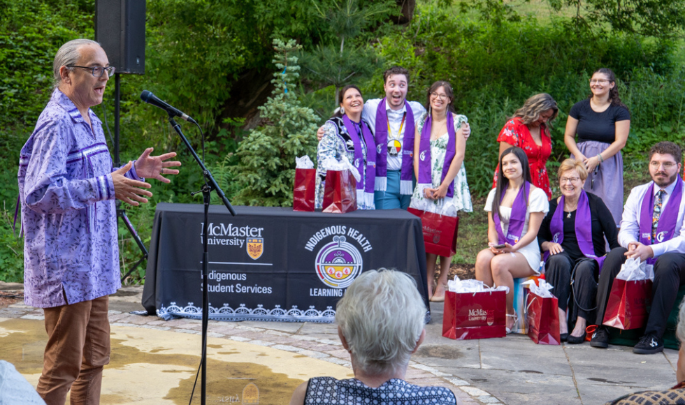 Rob Innes speaks at the mic at the outdoor classroom, watched by graduating students. 