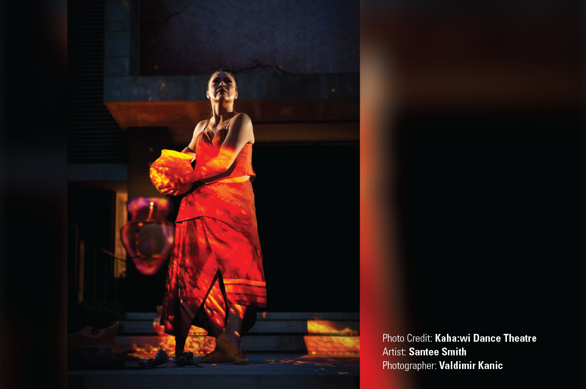 A photo of Santee Smith holding a ceramic pot with an orange light shining on her. 