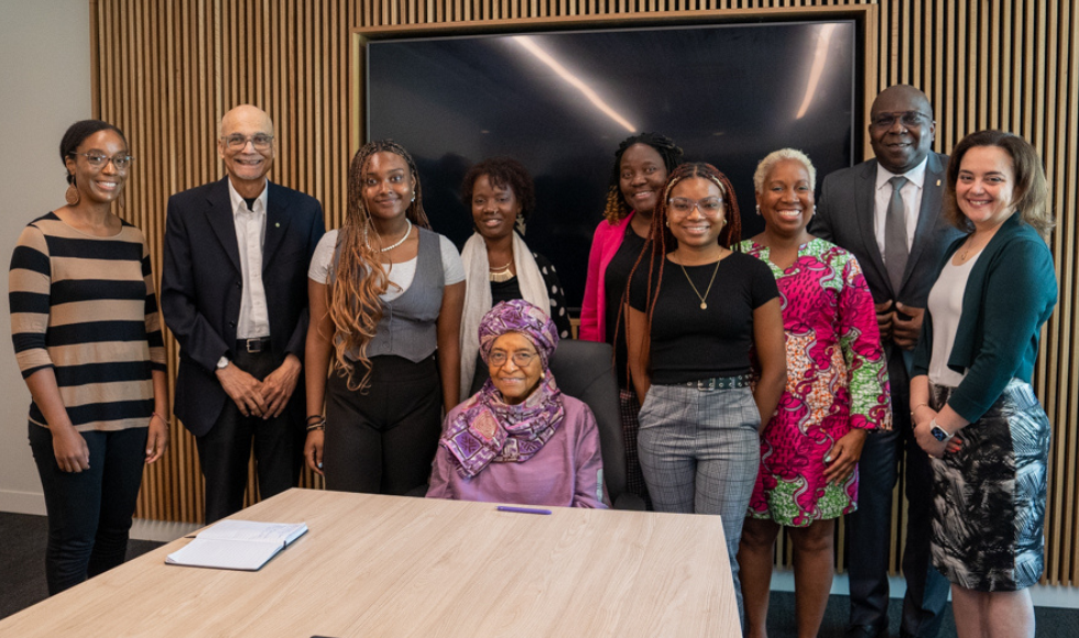 A group photo in a conference room with Sirleaf seated, flanked by a group of faculty members and two students