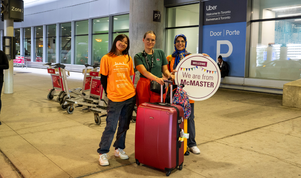 An arriving undergraduate student and student Airport Welcome Program volunteers smile at Pearson airport, standing with luggage.