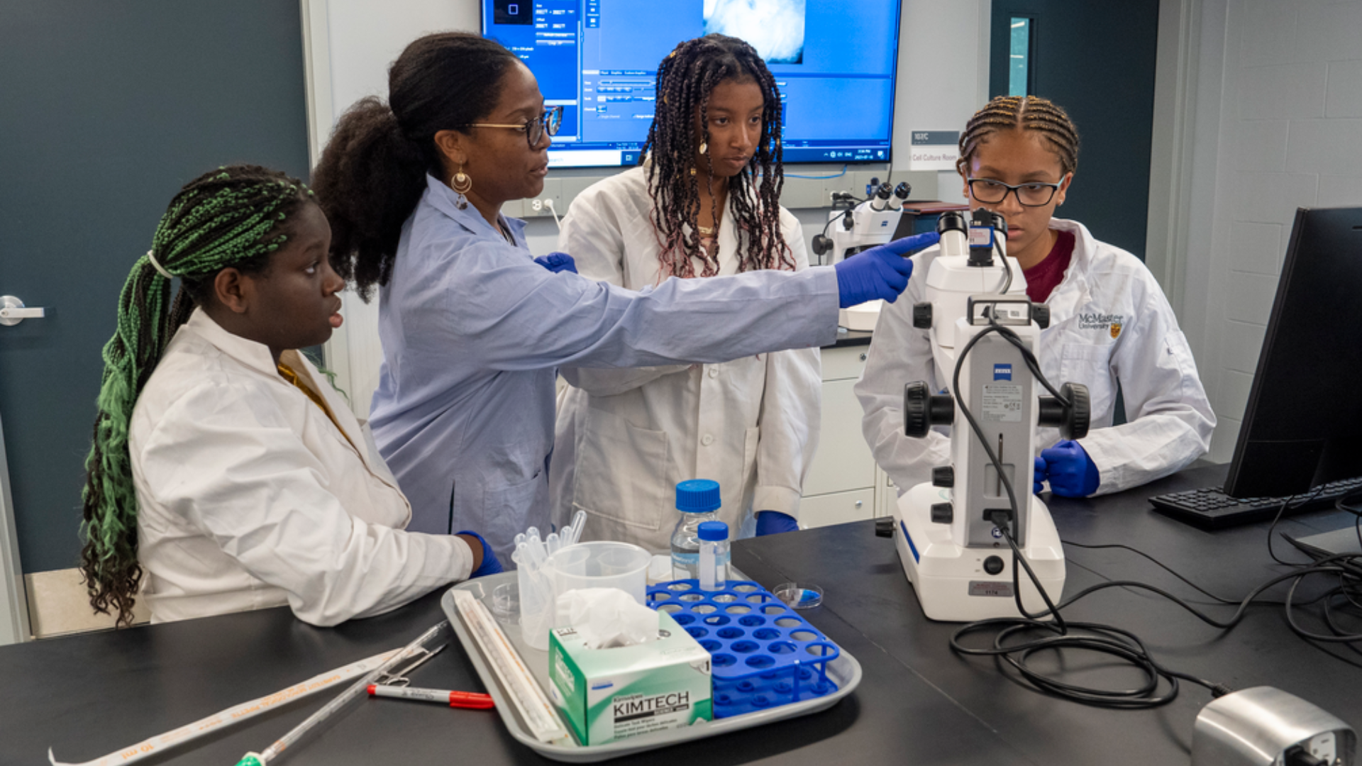 A professor and three students wearing lab coats conducting an experiement and working with a microscope 