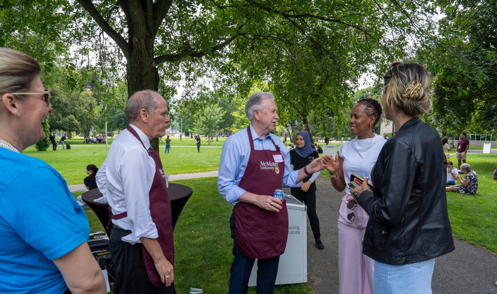 David Farrar and Paul O'Byrne, both wearing maroon aprons, laugh and chat with others at the employee picnic.