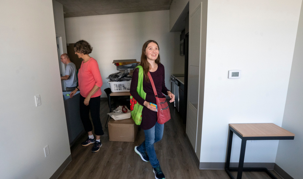  A young woman walks into an apartment. Behind her, there are moving boxes and two people unpacking. 