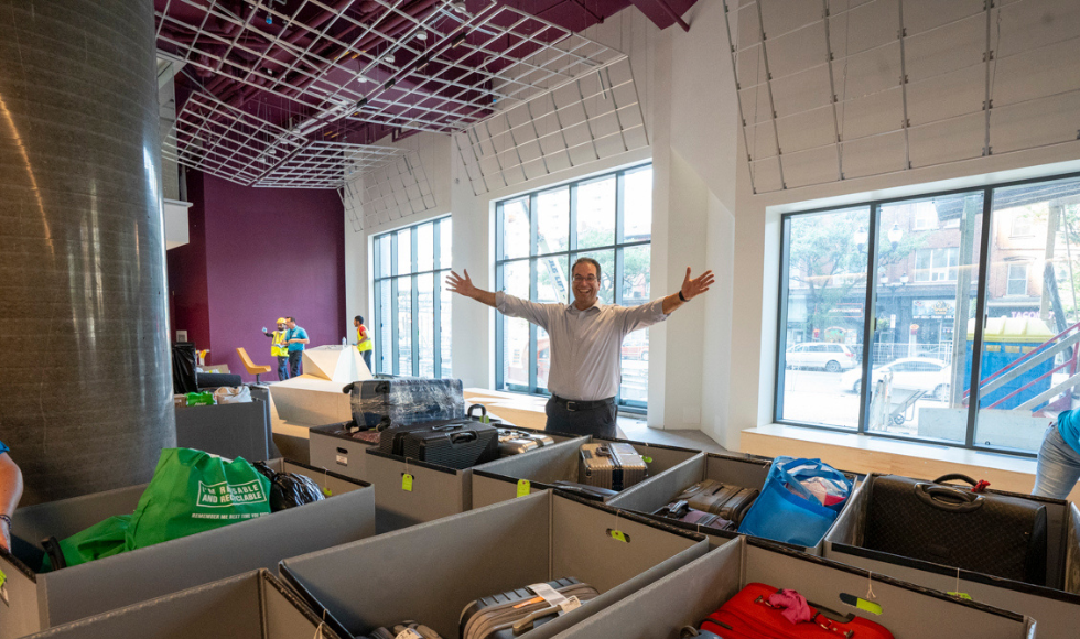 A smiling man with his arms outstretched with large boxes containing suitcases in front of him 