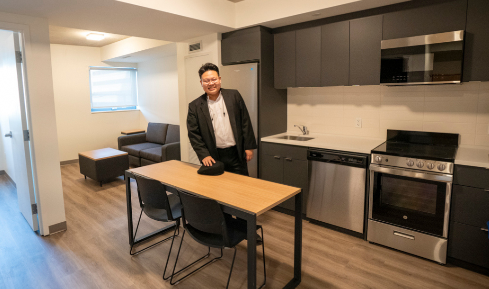 A student standing in the middle of an apartment smiles at the camera. 