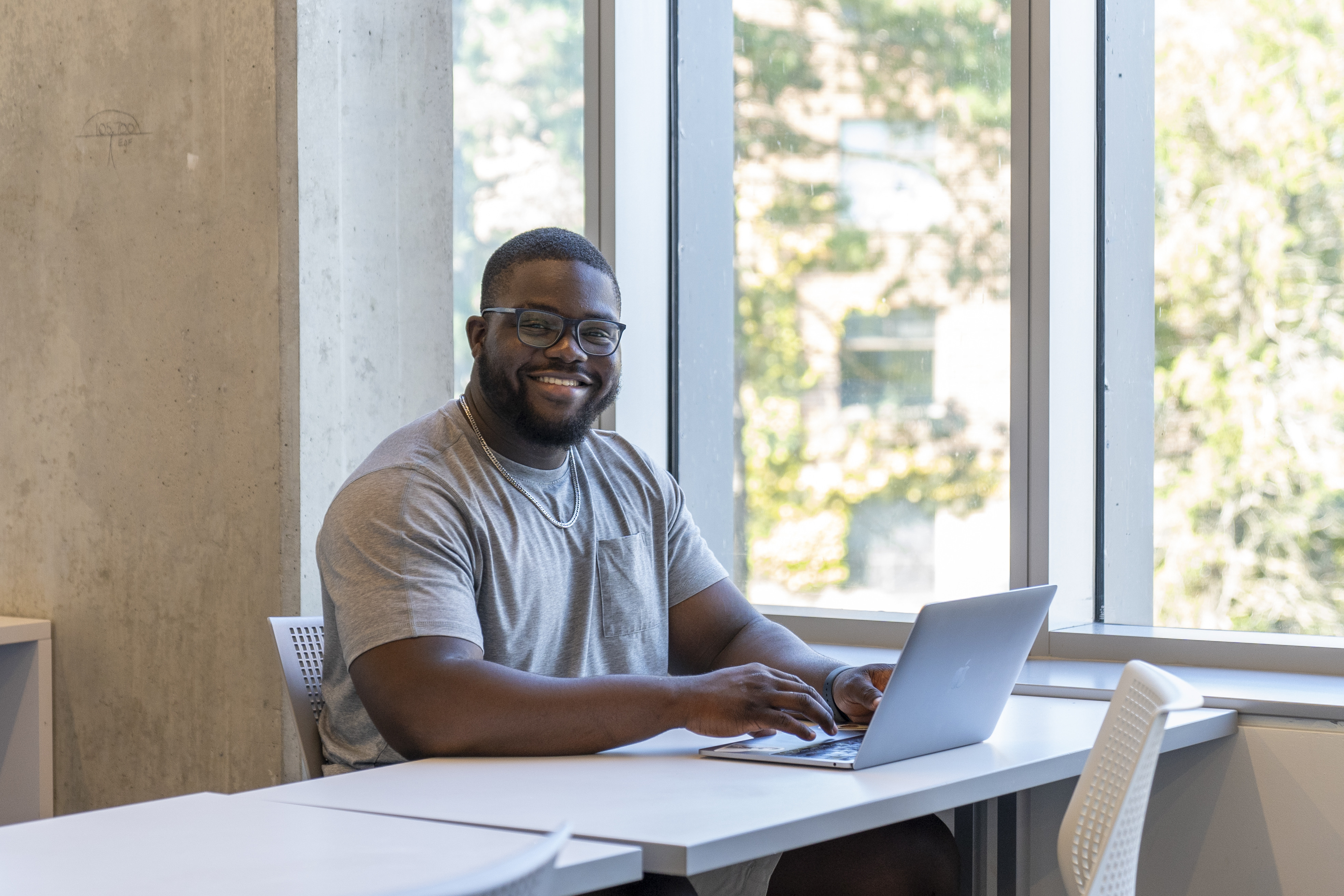 A student seated at a table with an open laptop in front of him. He is smiling at the camera. 