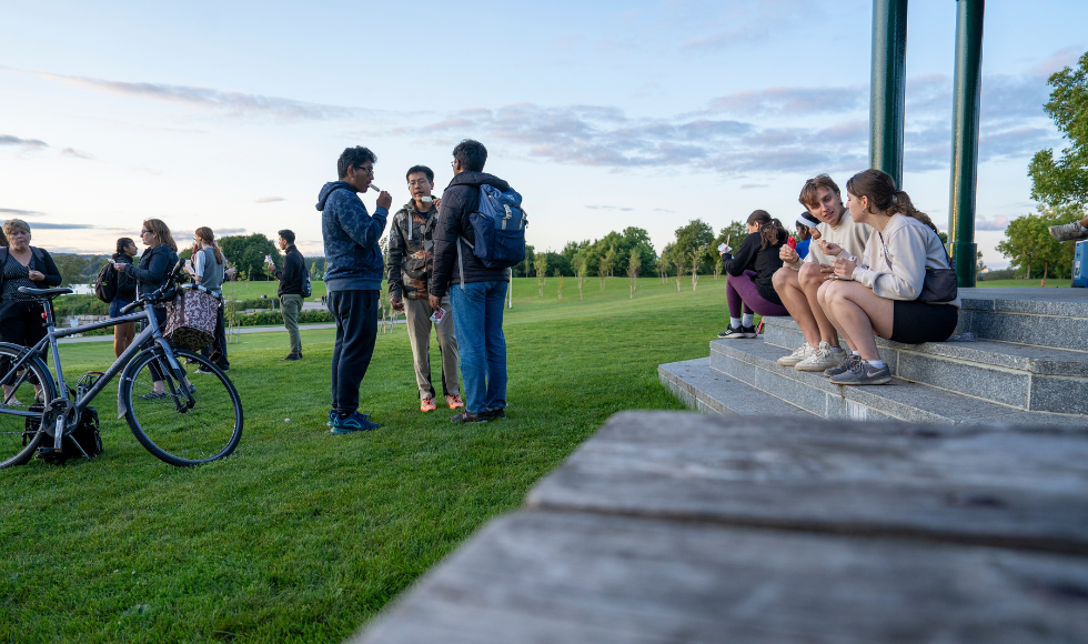 Students, some seated and some standing, eat frozen treats in a park 