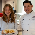 Two people, one holding a plate of food, and the other wearing chef's whites, smiling at the camera 