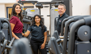 Three people standing surrounded by exercise equipment and smiling at the camera 