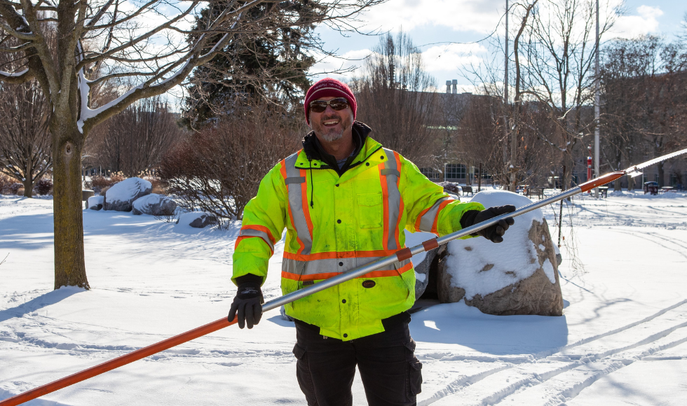 a facility services worker wearing a yellow vest holds a long pole while standing outdoors on campus on a bright snowy day