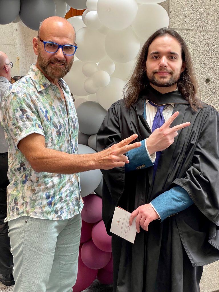 Two smiling men making the v for victory sign, one dressed in graduation gown.