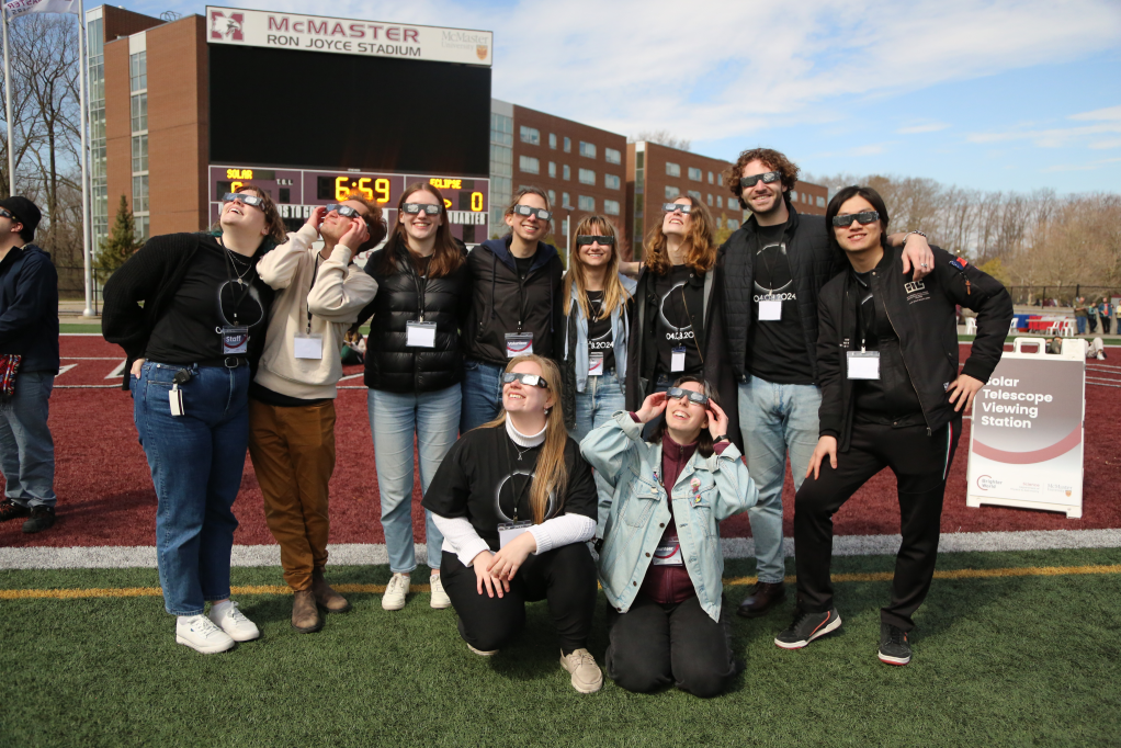 A smiling group of student eclipse ambassadors wearing their eclipse glasses, standing on the football field.