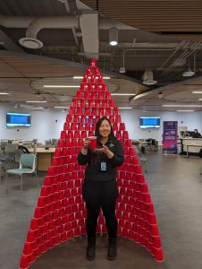A woman holding a stack of plastic red cups while standing in front of a rounded tower made up of dozens of plastic red cups. 