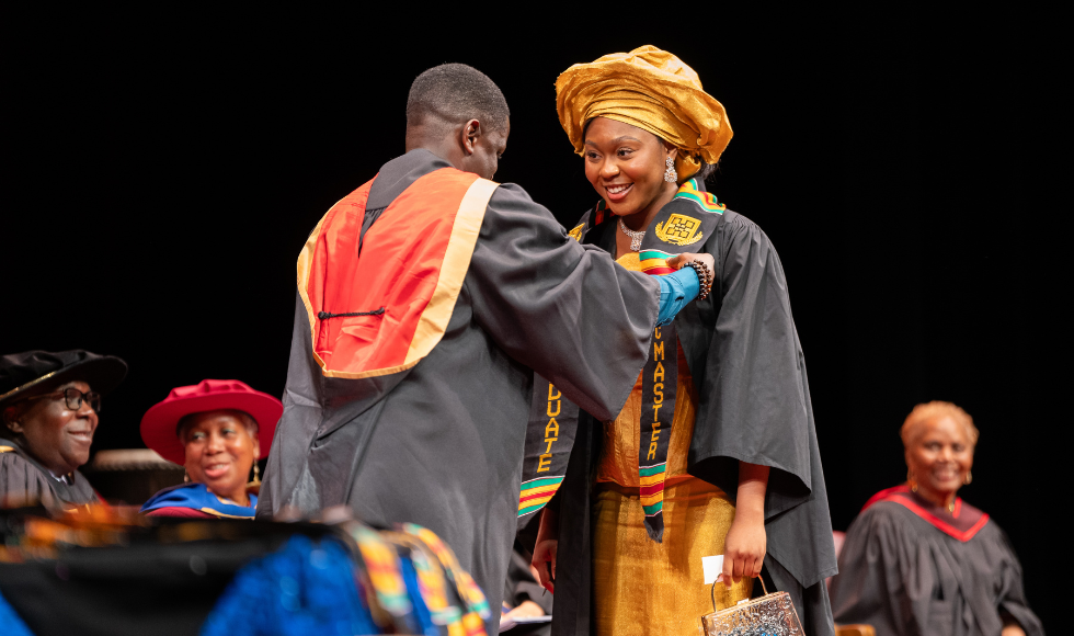 A woman receives her Kente stole from Mac Grad Leo Johnson on stage at the 2024 Black Excellence Graduation Celebration.