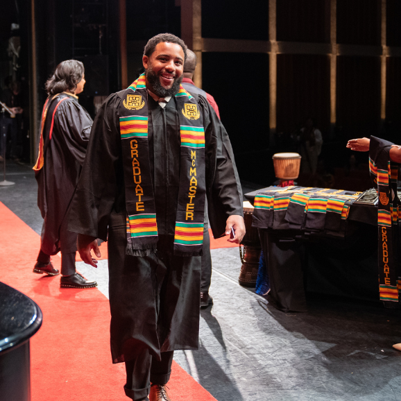 A man wearing a graduation gown and Kente Stole crosses the stage at the 2024 Black Excellence Graduation Celebration.