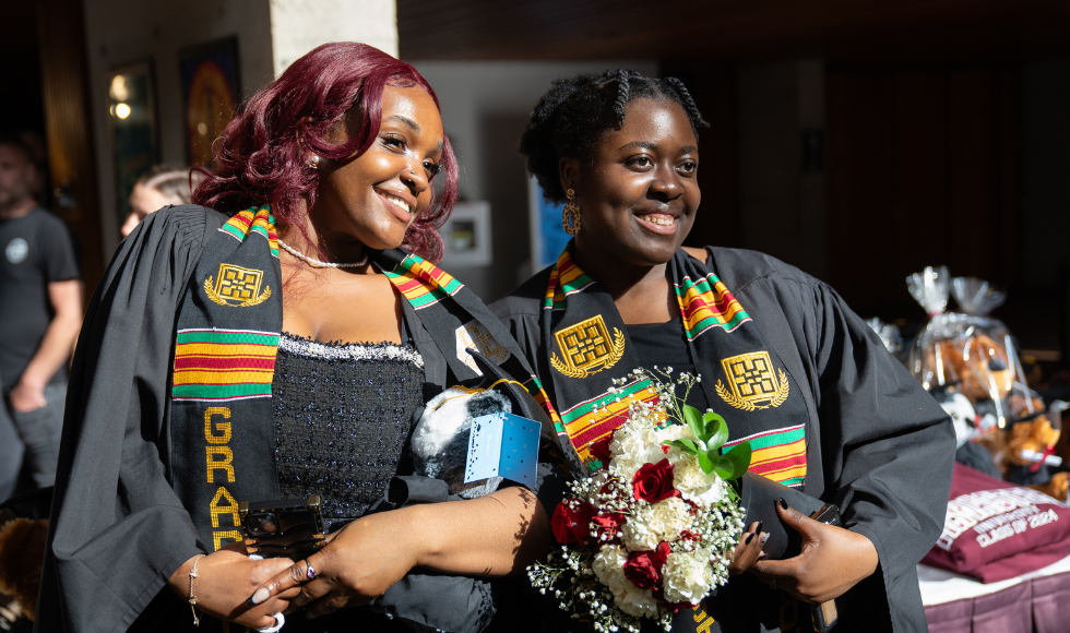 Two students in gowns and Kente stoles pose with flowers.