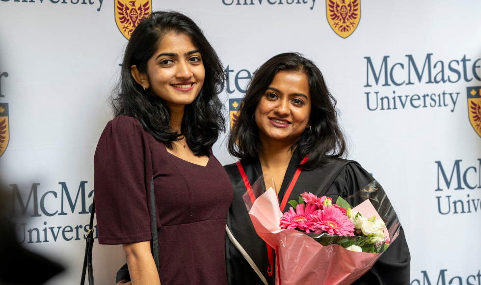 Two people, one wearing a graduation gown and holding a bouquet of flowers, smile for a photo 