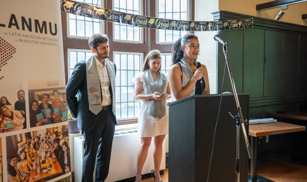 One person speaking into a microphone at a podium. Two other people are standing behind them.