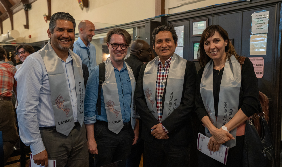 Four people standing and smiling at the camera. They all have grey convocation stoles around their shoulders.