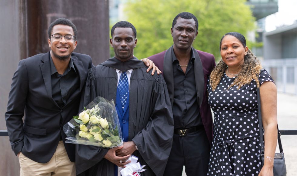 Four people, one of whom is wearing a graduation gown and holding a bouquet of flowers, pose for a photo