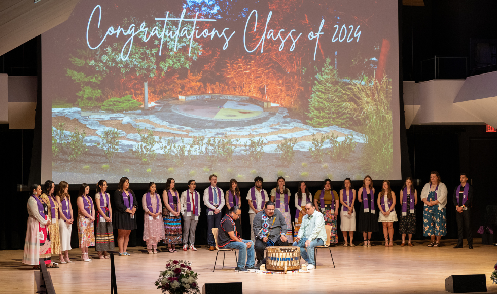 A line of people wearing purple stoles look on as three people participate in an Indigenous drumming circle. Behind them is a screen that reads, 'Congratulations Class of 2024.' 