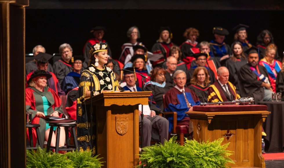 A woman in ceremonial convocation regalia speaking at a podium. Behind her is a crowd of people wearing colourful convocation gowns. 