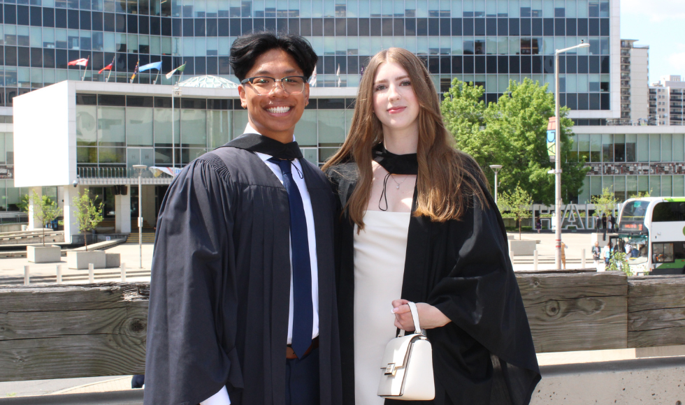 Two people in graduation robes standing outdoors smiling at the camera 