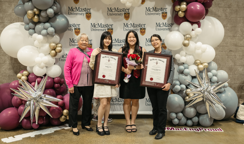 Four people standing in front of a McMaster-branded backdrop. Two of them are holding framed degrees, while another is holding a bouquet of flowers 