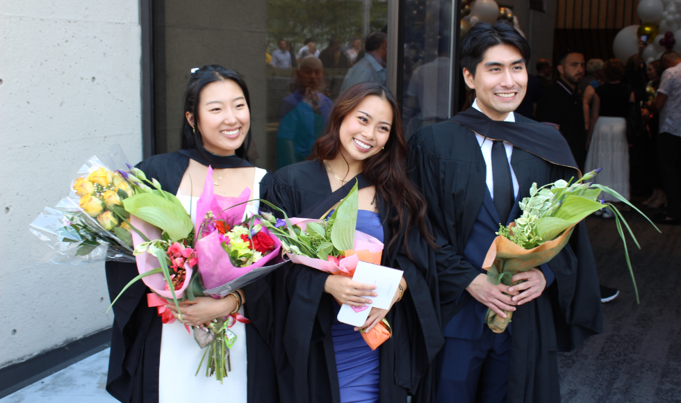 Three people in graduation robes, each smiling and holding a bouquet of flowers 