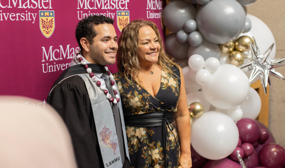 Two people, one of whom is wearing a graduation gown and stole, smile and post for a photo against a McMaster-branded backdrop