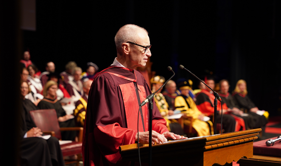 A man in a maroon and red graduation gown speaks into a microphone at a podium 