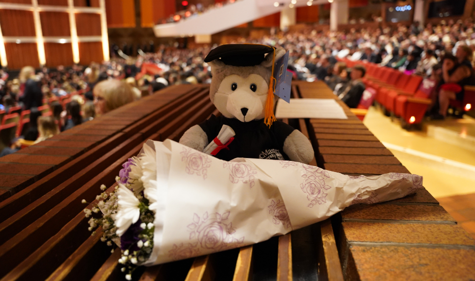 A teddy bear that has a convocation cap and gown set on a surface with a bouquet of flowers in front of it