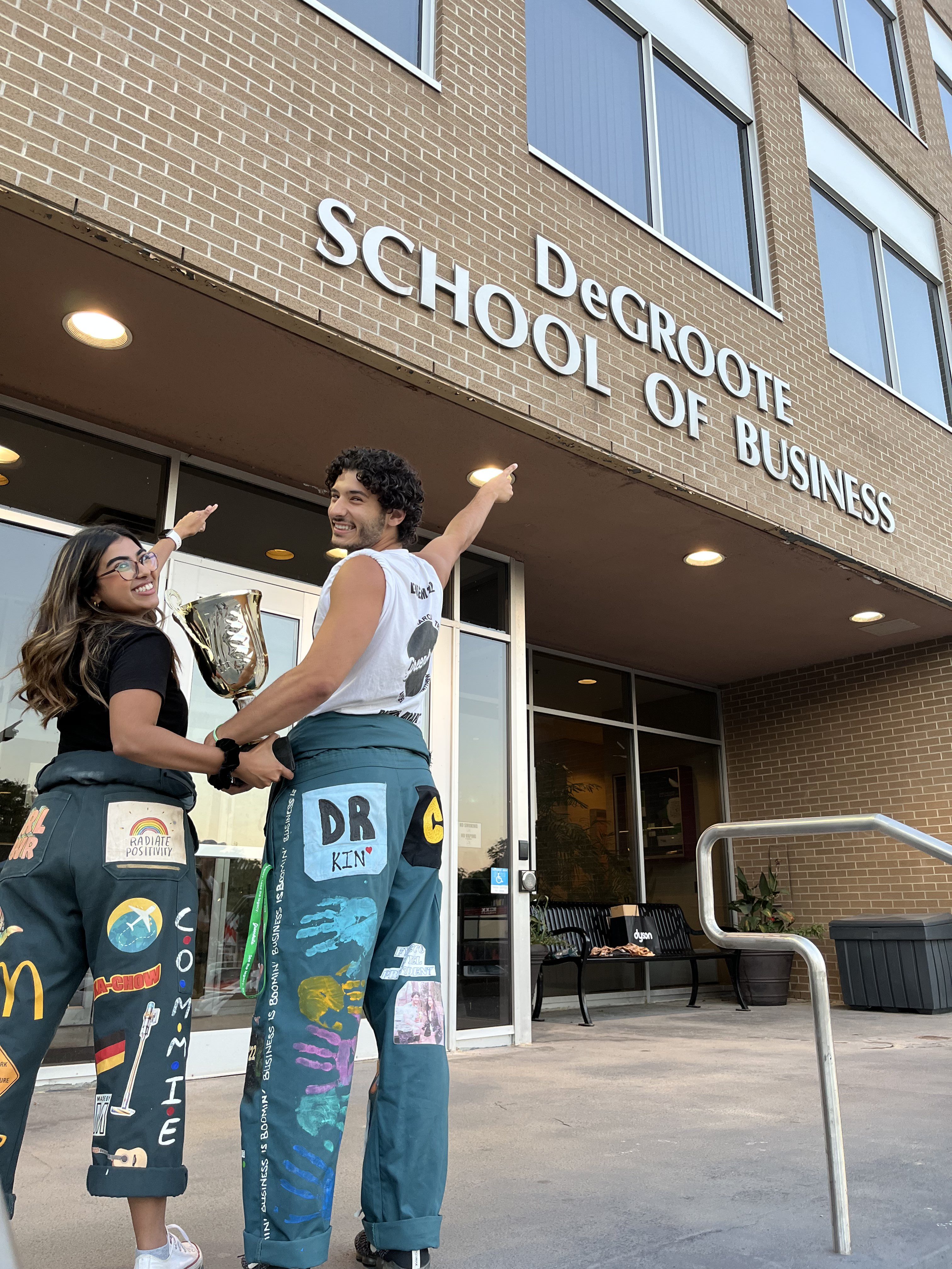 Two McMaster students in Welcome Week apparel holding a trophy and pointing to a sign on the exterior of a building that reads, 'DeGroote School of Business.' 