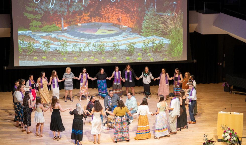 A large group of people holding hands in a big ring around 3 traditional drummers on stage.