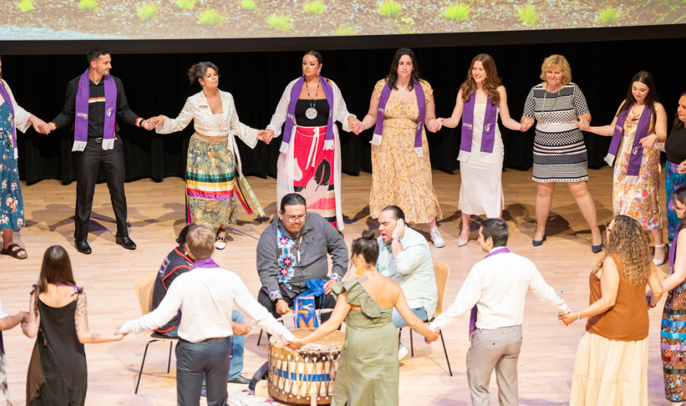 A large group of people holding hands in a big ring around 3 traditional drummers on stage.