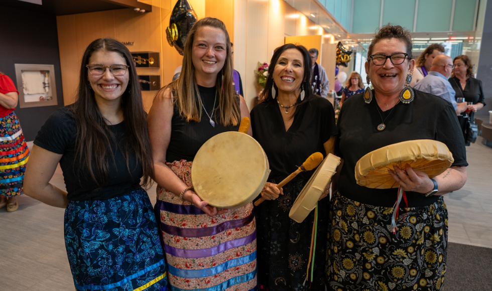 Four smiling women holding hand-held drums (one behind her back) at the Indigenous grad celebration.