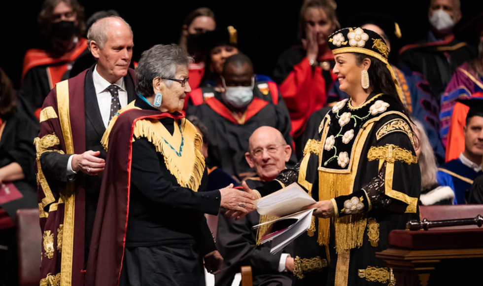 Bertha Skye on stage shaking the hand of McMaster Chancellor Santee Smith while McMaster President David Farrar. There are people seated in the background and everyone is dressed in convocation regalia. 