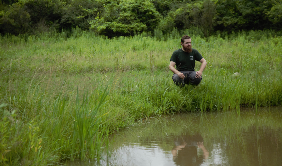 Noah Stegman crouched down in tall grasses looking at a body of water.