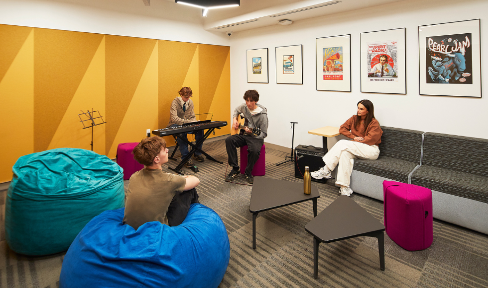 Four people seated in a room. One is playing an electric piano and one is playing an acoustic guitar while the other two appear to be listening.