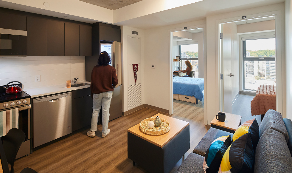 The interior of a two bedroom apartment. One person can be seen in the kitchen and another can be seen seated at a desk in one of the bedrooms.