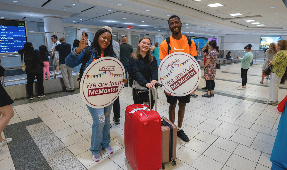 An smiling international student with a suitcase stands at the airport with two people from McMaster's Airport Welcome program. 