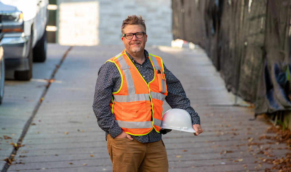 Smiling Marc St-Jean in a reflective vest, holding his hard hat against a backdrop of construction.